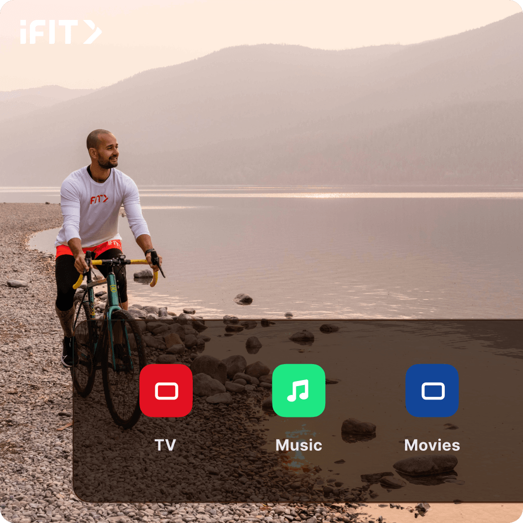 iFIT Trainer Paulo Barreto rides a green and yellow bike along a pebbled beach with a foggy lake and mountains in the background.